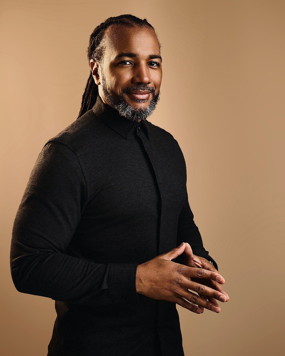 Usama Young, standing in a charcoal shirt, hands together, smiling at the camera